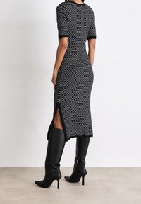 Kobieta w midi sukience w czarno-białe wzory z bocznym rozcięciem oraz czarnych wysokich kozakach do kolan, stojąca na jasnej podłodze.