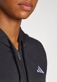 Black zip-up hoodie with a high collar, featuring a textured fabric, subtle gray logo on the left chest, and a smooth zipper.