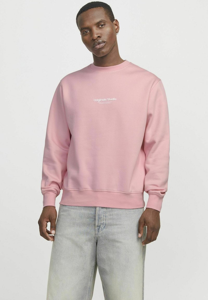 Hele roosa crewneck sweatshirte, mis on valmistatud pehmest kangast, millel on rinnal valge "Originals Studio" logo ja soonikkoes varrukad.