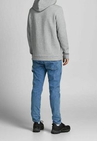 Felpa grigia con cappuccio, jeans blu con tasca laterale e scarpe nere. Design semplice e vestibilità casual, vista da dietro.