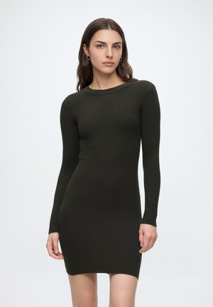 Femme aux longs cheveux bruns portant une robe moulante côtelée à manches longues vert foncé, debout devant un fond gris clair uni.