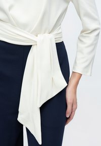Hvit, myk stofftopp med en lang, knyttet midjebelte, kombinert med mørkeblå bukser. Toppen har lange ermer og en avslappet passform.