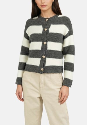 Femme portant un cardigan boutonné à larges rayures grises et blanches et un pantalon beige, debout les bras détendus le long du corps.