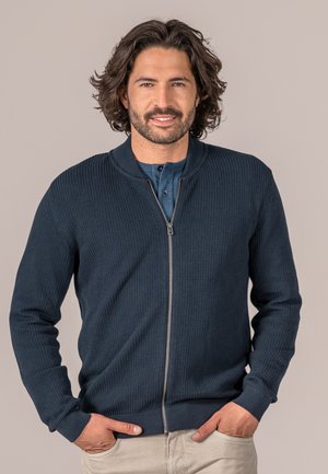 Marineblaues Reißverschluss-Cardigan mit strukturiertem Muster, geripptem Kragen und Bündchen sowie einem glatten Reißverschluss. Über einem blauen Hemd mit Knöpfen getragen.