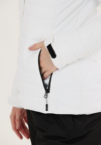 Hvid oppustelig jakke med en sidelukket lomme og sorte detaljer ved kraven og manchetterne. Glat tekstur med syede vandrette linjer.