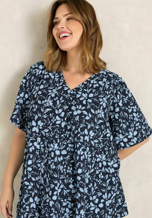 Kobieta uśmiechająca się, ubrana w luźną czarną bluzkę z jasnoniebieskimi kwiatowymi wzorami i krótkimi rękawami na jednolitym beżowym tle.