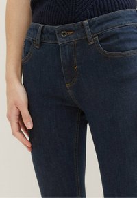 Tummansiniset denim-farkut, joissa on istuva malli, kullanväriset tikkausompeleet, etutaskut ja vyötäröllä nappikiinnitys.