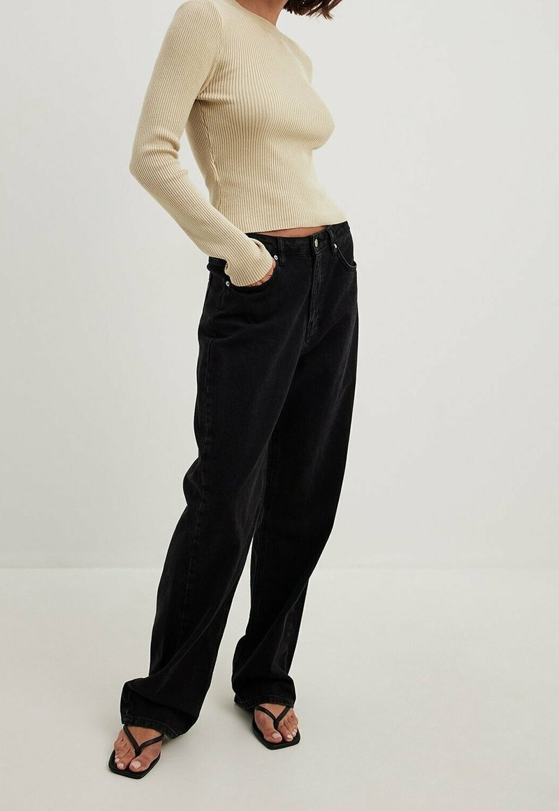 Jean noir taille haute avec une jambe droite, présentant des passants de ceinture et des poches avant. Associé à un haut à manches longues beige côtelé.