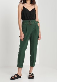 Camisole en satin noir avec dentelle et boutons, associée à un pantalon taille haute vert avec une ceinture et des ourlets retroussés, et des sandales noires ornées de embellissements.