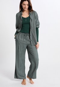 Vrouw staat blootsvoets en draagt een donkergroene longsleeve met een bijpassende groen-witte geruite blouse en losse broeken.
