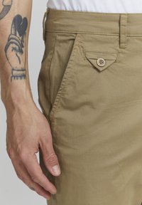 Beige lightweight trousers with a textured finish, featuring a triangular pocket detail with a button and visible stitching on the waistband.