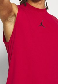 Červené bezrukávové sportovní tričko vyrobené z bavlny, s kulatým výstřihem a černým logem Jordan na hrudi. Hladká textura a volný střih.