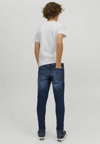 Vit kortärmad t-shirt, blå jeans med blekning, synliga fickor, figurnära design och svarta sneakers. Vy från baksidan.