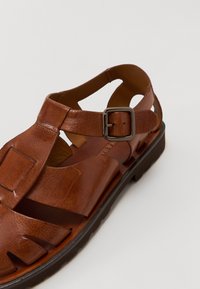 Brune skinnsandaler med utskjæringsdesign, justerbar ankelrem med sølvspenne og teksturert gummisåle.