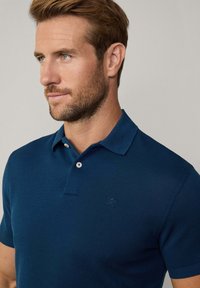 Marinblå pikétröja med texturerad tyg, två vita knappar och en diskret präglad logotyp på bröstet. Korta ärmar och klassisk krage.