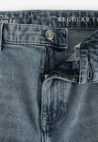 Jeans din denim cu nuanță albastră, cu fermoar și închidere cu buton din metal. Tivuri cusute și un design cu potrivire obișnuită, având o textură tipică de uzură.