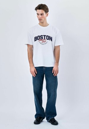 Ung mand stående iført en hvid "Boston Athletics Dept" t-shirt, blå jeans og sorte sneakers mod en ensartet lys baggrund.