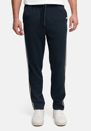Uomo con pantaloni neri da ginnastica con strisce laterali bianche e sneakers bianche, in piedi contro uno sfondo semplice.