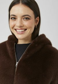 Sweat à capuche marron en fausse fourrure avec une texture douce, doté d'une grande capuche, d'une fermeture éclair argentée, porté sur un haut noir.