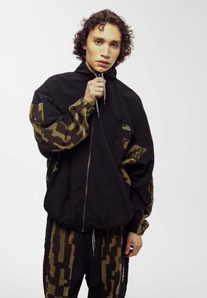 Schwarze Windbreaker-Jacke mit camouflagemusternden Ärmeln, Frontreißverschluss und Kordelzug am Kragen. Getragen mit passenden gemusterten Hosen.