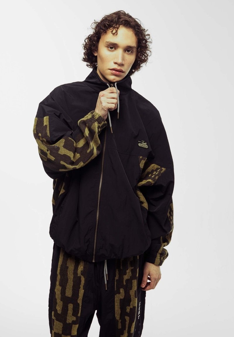 Chaqueta cortavientos negra con mangas de estampado de camuflaje, cremallera frontal y cuello con cordón. Lleva pantalones con el mismo estampado.
