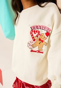 White sweatshirt featuring a sewn gingerbread man graphic, "FESTIVE" text, and a candy cane design. Ribbed cuffs and relaxed fit.
