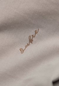 Tissu beige clair avec de petits fils de broderie bruns délicats en écriture cursive formant un mot près du centre.