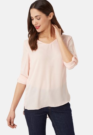 Mujer con cabello castaño a la altura de los hombros, vestida con una blusa rosa claro y jeans azul oscuro, tocándose el cuello y mirando hacia abajo, sonriendo.