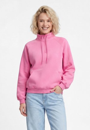 Smilende kvinde med blondt hår iført en pink pullover med halv lynlås og lyseblå jeans, hånd i lommen, stående mod en hvid baggrund.