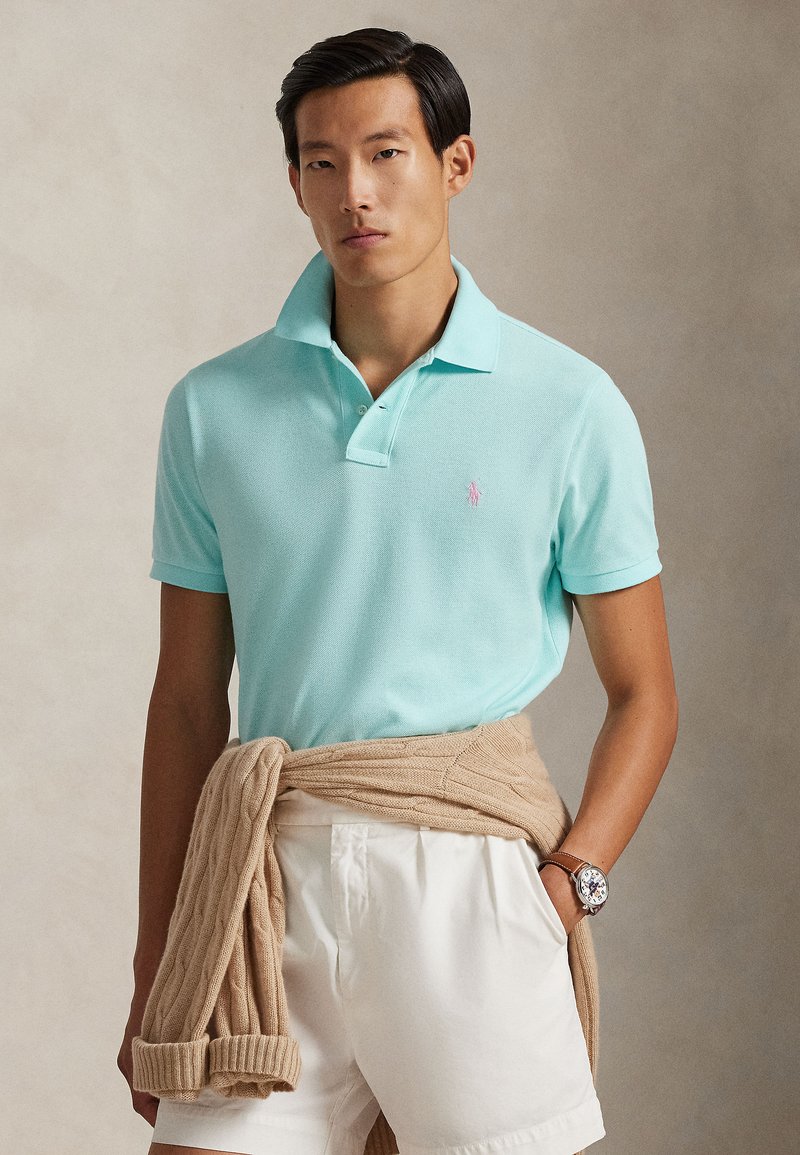 Ανοιχτό γαλάζιο κοντομάνικο πόλο με πλεκτή υφή και μικρό ροζ λογότυπο. Μπεζ πουλόβερ δεμένο γύρω από τη μέση, λευκό σορτς, ρολόι χειρός.