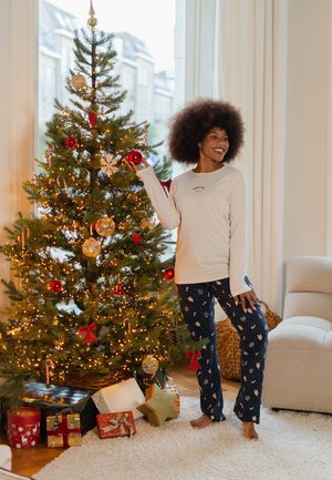 Crèmekleurig shirt met lange mouwen en logo, gecombineerd met marineblauwe broek met bloemenpatroon. Op de achtergrond is een versierde kerstboom en cadeaus te zien.