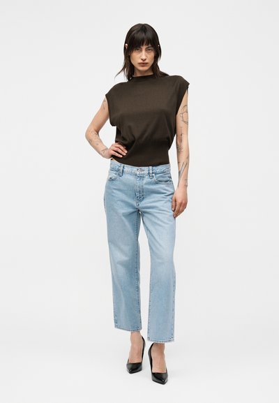 Γυναίκα με τατουάζ φοράει μαύρο αμάνικο τοπ, ανοιχτό μπλε τζιν με ίσια γραμμή και μαύρες ψηλές γόβες, στέκεται μπροστά σε έναν απλό φόντο.