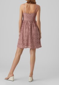 Vestido de encaje en rosa empolvado con tirantes finos, un cuerpo ajustado, falda acampanada y un patrón texturizado. Combinado con sandalias de cuña.