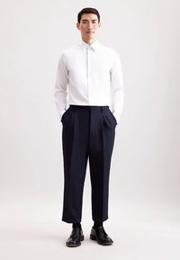 Camicia bianca con bottoni, pantaloni plissettati blu navy, scarpe nere. Vestibilità aderente, tessuto liscio, colletto netto, design su misura.