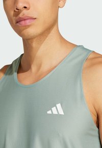 Svijetlozelena sportska majica bez rukava s teksturiranom tkaninom i okruglim izrezom. Ima bijeli logo s tri dijagonalne pruge na lijevoj strani prsnog koša.