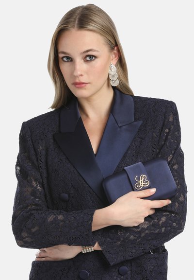 Kvinde med blondt hår iført en mørk blondeblazer, der holder en marineblå clutch med guldlogo, accessoriseret med store udsmykkede øreringe og et armbånd.