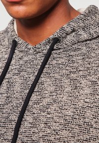 Harmaa ja musta neulottu huppari, jossa on teksturoitu kuvio. Hupparissa on mustilla yksityiskohdilla varustettu naru ja tiukasti istuva huppu.