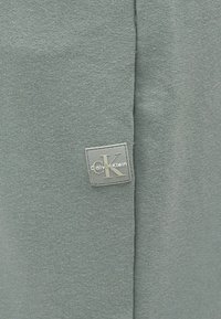 Μαλακό γκρι-πράσινο πλεκτό ύφασμα με μικρή τετράγωνη ετικέτα που φέρει το λογότυπο "CK" και "Calvin Klein" κοντά σε μια ραφή.