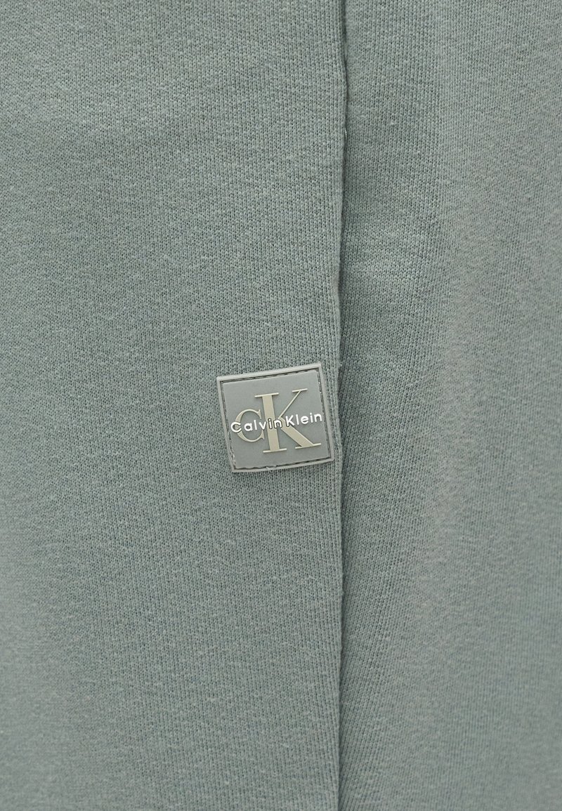 Tissu tricoté doux gris-vert avec une petite étiquette carrée comportant le logo "CK" et "Calvin Klein" près d'une couture.