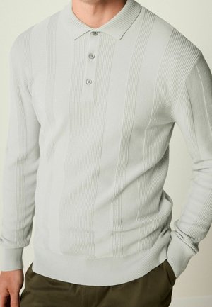 Mann trägt hellgrauen, langärmligen, gestrickten Polo-Pullover mit vertikalen strukturierten Streifen und olivgrüner Hose, Hand in der Tasche.
