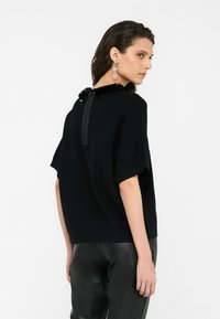 Black short-sleeve top with textured floral accents at the neckline and a central zipper detail at the back, paired with shiny black pants.