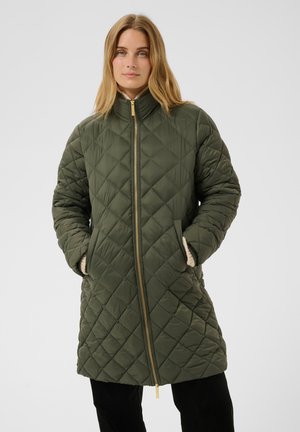 Femme portant une veste matelassée verte longueur genou avec une fermeture éclair dorée et les mains dans les poches, debout devant un fond uni.