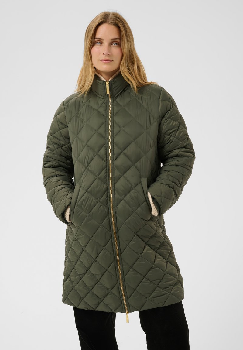 Femme portant une veste matelassée verte longueur genou avec une fermeture éclair dorée et les mains dans les poches, debout devant un fond uni.