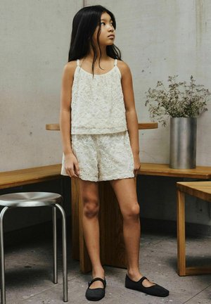 Pige med langt sort hår iført en hvid blonde-top og shorts står ved træborde og en metal skammel i et minimalistisk rum.