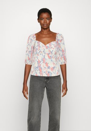 Blusa com padrão floral, com decote quadrado, mangas bufantes, em tons pastel incluindo rosa e azul, combinada com jeans cinzentos de cintura alta.