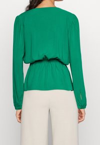 Blusa verde a maniche lunghe con vita arricciata, scollatura tonda e dettagli plissettati sulle spalle. Texture liscia e tessuto leggero.