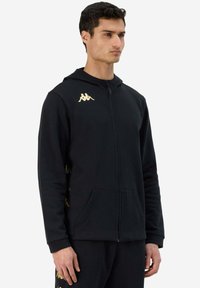 Felpa nera con zip realizzata in tessuto morbido, con un logo dorato sul petto e dettagli gialli lungo le maniche. Includi tasche frontali.