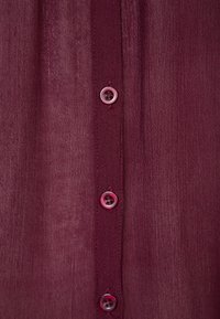 Chemise en tissu bordeaux avec une patte de boutonnage comportant quatre boutons rose clair et une finition douce et texturée.