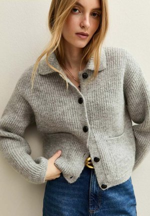 Young woman wearing a grey ribbed button-up cardigan, blue jeans, and gold belt buckle, posing with one hand in pocket against neutral background.