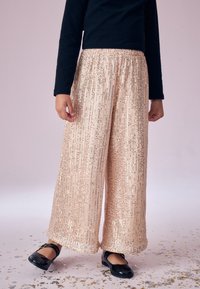 Pantaloni a gamba larga di un delicato rosa pallido, ricoperti di paillettes e con una texture morbida, dotati di una fascia elastica in vita, abbinati a scarpe nere in vernice.